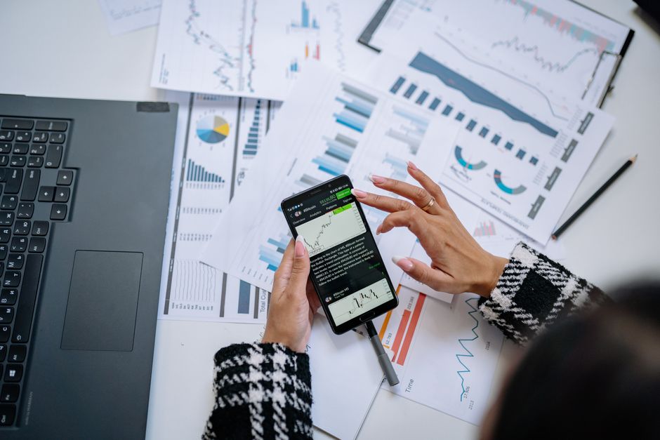 Businesswoman analyzing financial charts on smartphone with laptop and documents.