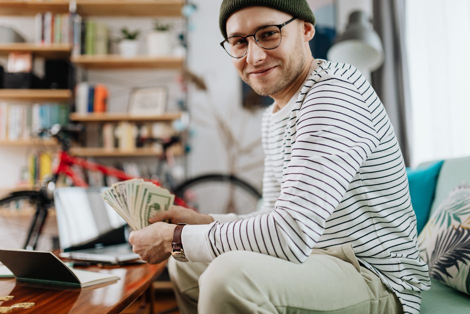 Smiling man with glasses counting cash, sitting in a cozy living room with a bicycle in the background.