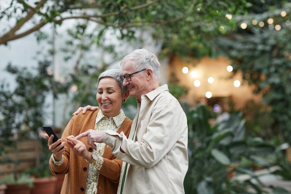 Happy senior couple using smartphone together in lush garden setting, embracing technology and leisure.
