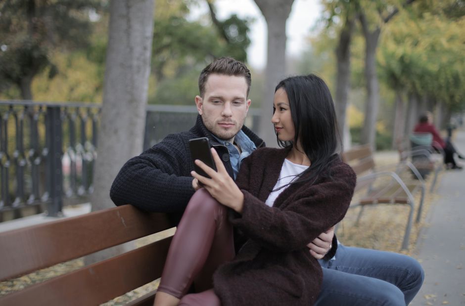 Loving modern multiracial couple in casual warm clothes sitting together on bench and using smartphone while spending time in park in city