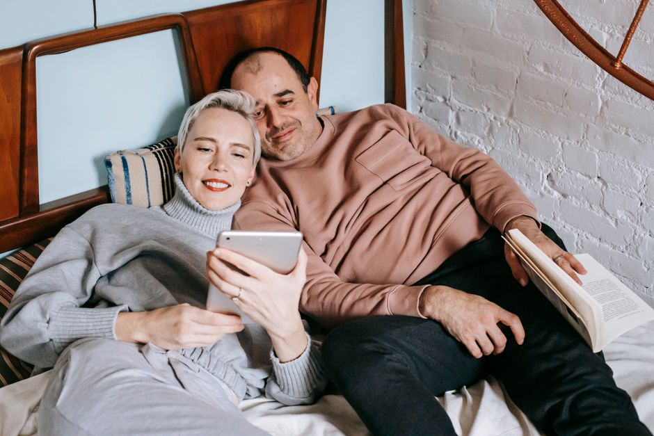 From above of happy adult diverse couple in casual outfits lying on bed and sharing table during weekend at home