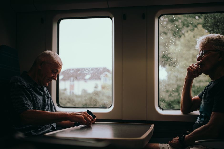 Two senior men travel by train in London, England, gazing out the window at the lush countryside.