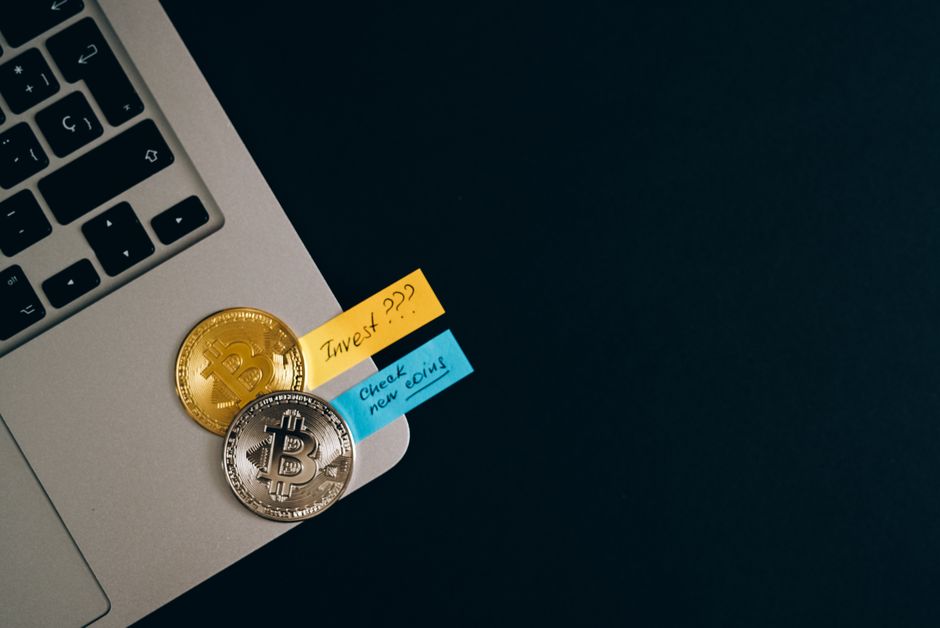 Bitcoin coins placed on a laptop keyboard with sticky note reminders about investing and checking new coins.