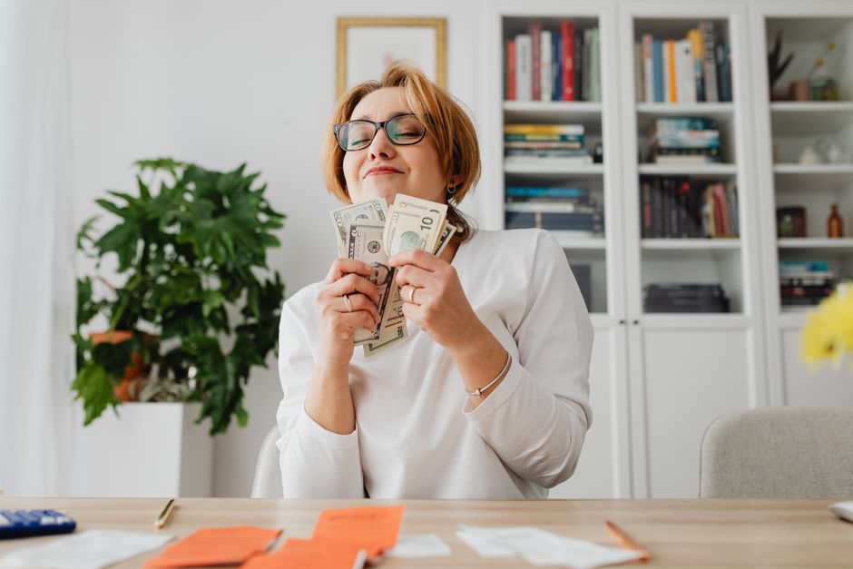 Smiling woman holding dollar bills at home desk surrounded by receipts and bookshelves.