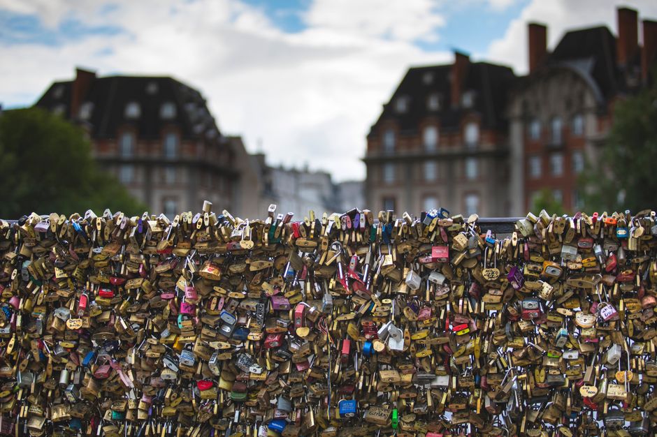 Close-up of love padlocks on a bridge with historic buildings in the background on a cloudy day.