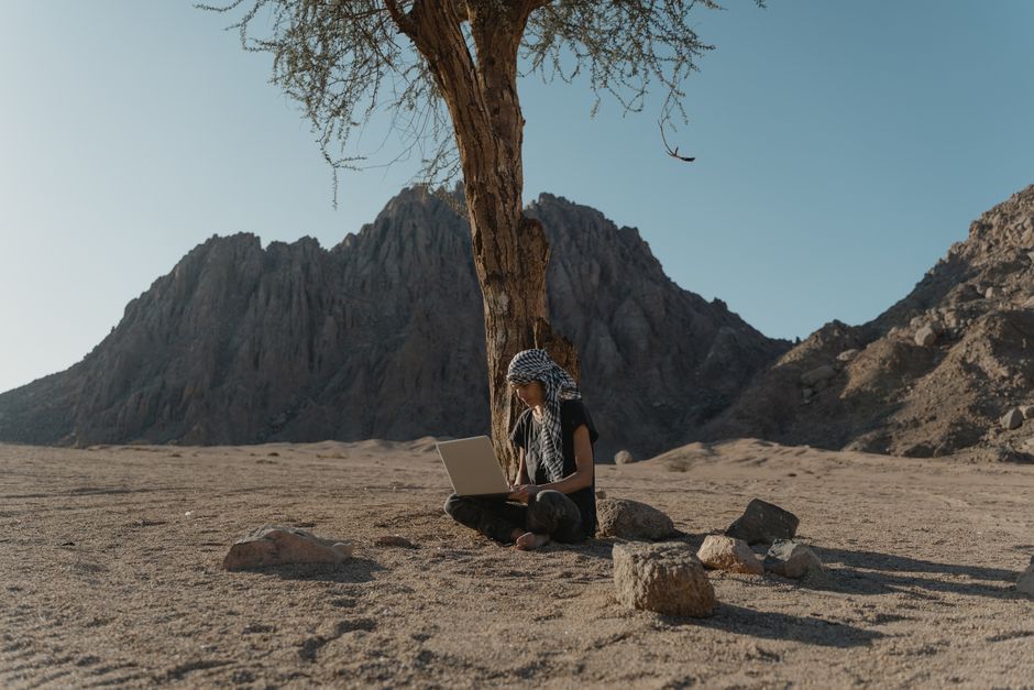 Caucasian woman using laptop under tree in rocky desert landscape, embracing remote work outdoors.